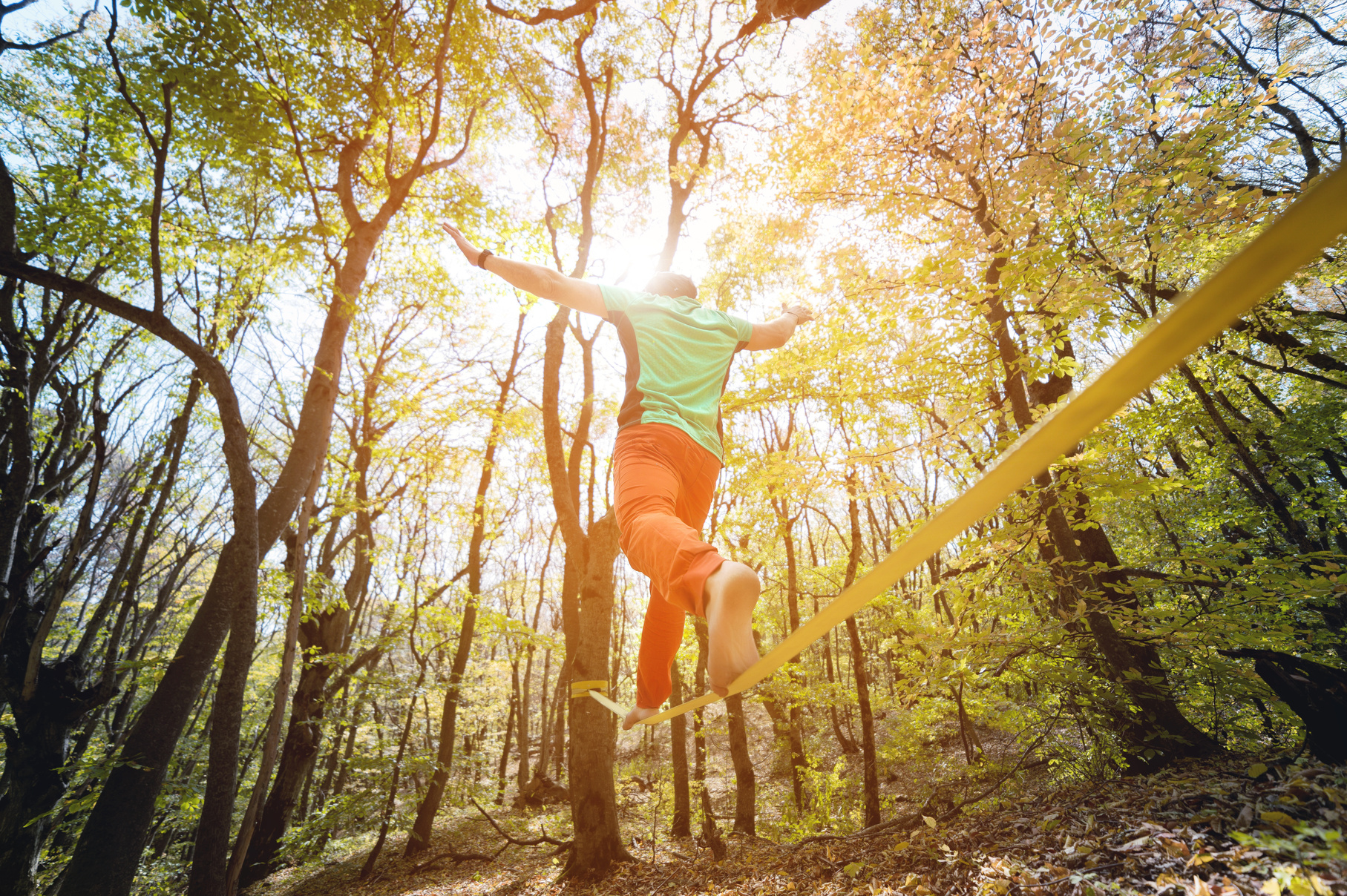 Wide angle male tightrope walker balancing barefoot on slackline in autumn forest.